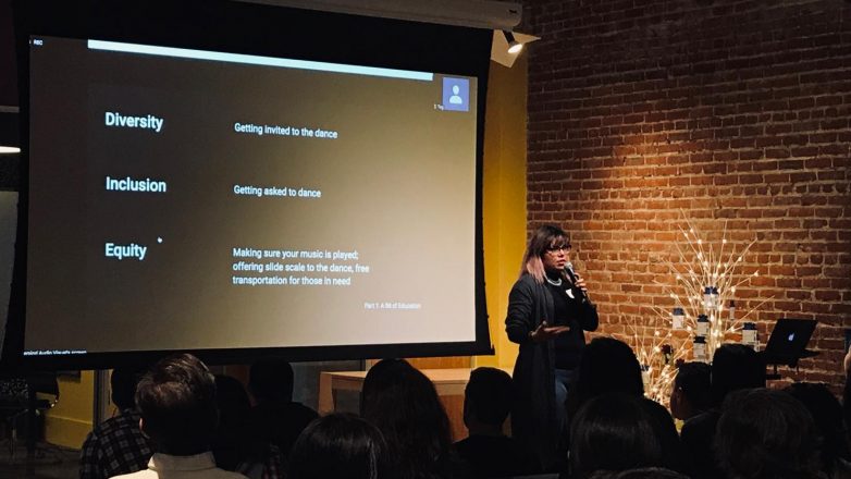 Alexis in the foreground. Behind her, a large screen displays her slide with the definitions of diversity, equity, and inclusion