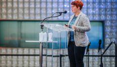 A woman with a red pixie haircut and orange glasses standing at a clear podium and speaking into a microphone. There is a lighted glass block wall behind her and blurry audience member heads in the foreground.