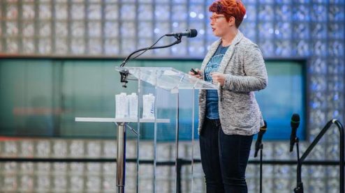 A woman with a red pixie haircut and orange glasses standing at a clear podium and speaking into a microphone. There is a lighted glass block wall behind her and blurry audience member heads in the foreground.