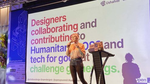 Eriol standing on stage in front of a slide that reads 'Designers collaborating and contributing to Humanitarian OSS and tech for good at challenge gatherings.'