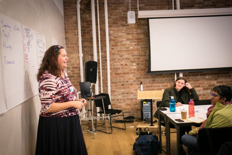 A woman standing at the front of a room in front of a whiteboard talking to people sitting at tables as they take notes.