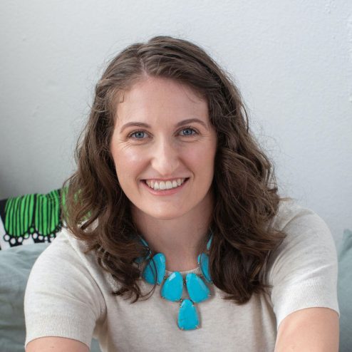 Woman with wavy brown hair wearing a beige shirt and large turquoise necklace smiling at the camera