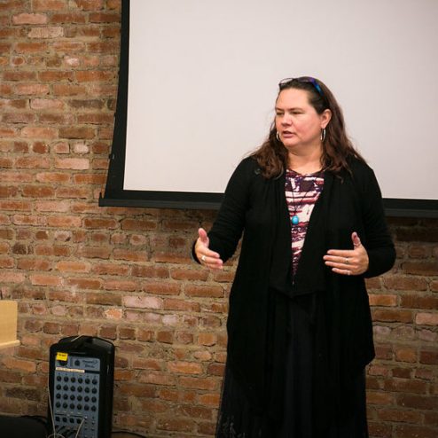 A woman wearing a black sweater, black skirt, and striped shirt, with dark brown hair. She is standing in front of a projection screen talking to an unseen audience and gesturing with her hands.