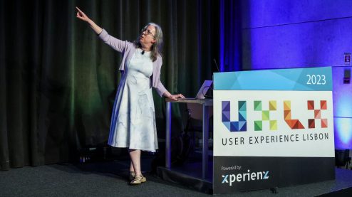 Woman in glasses and a white dress points toward the top left standing next to a poster that says UXLX in colorful print