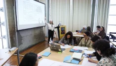 Classroom with several people at tables watching a woman in black pants and a white shirt gesturing at a large screen