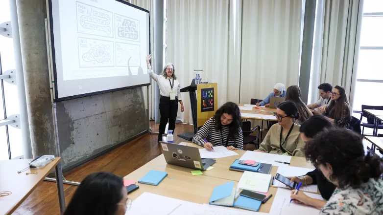 Classroom with several people at tables watching a woman in black pants and a white shirt gesturing at a large screen